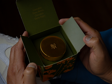 Hand holding a small round container of Texas grown hemp with a Hye Harvest lid, inside an open box, against a dark background.