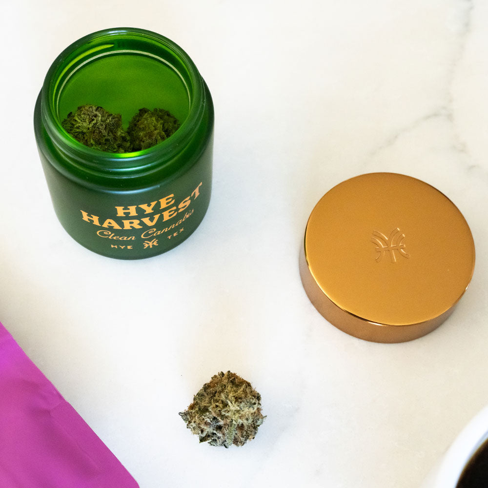 A frosted green glass jar of Hye Harvest Wild Flower sitting on a marble surface next to a gold lid and a cup of coffee. This clean-burning botanical is designed for a bright daytime lift and clear focus during a morning creative ritual.