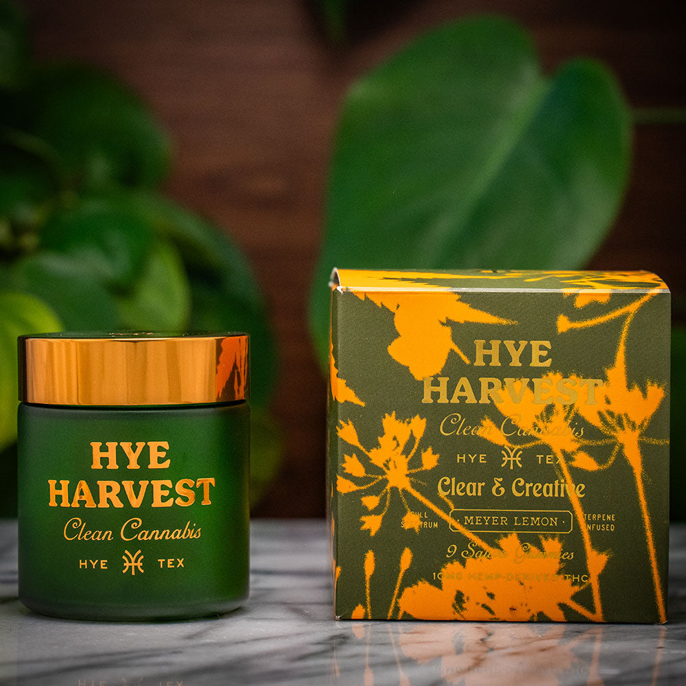 A green glass jar with a gold lid sits next to its olive green and yellow patterned packaging box on a marble countertop, with both items featuring gold lettering that reads "Hye Harvest Clean Cannabis Clear & Creative Meyer Lemon Sativa Gummies" in front of a soft green plant background.
