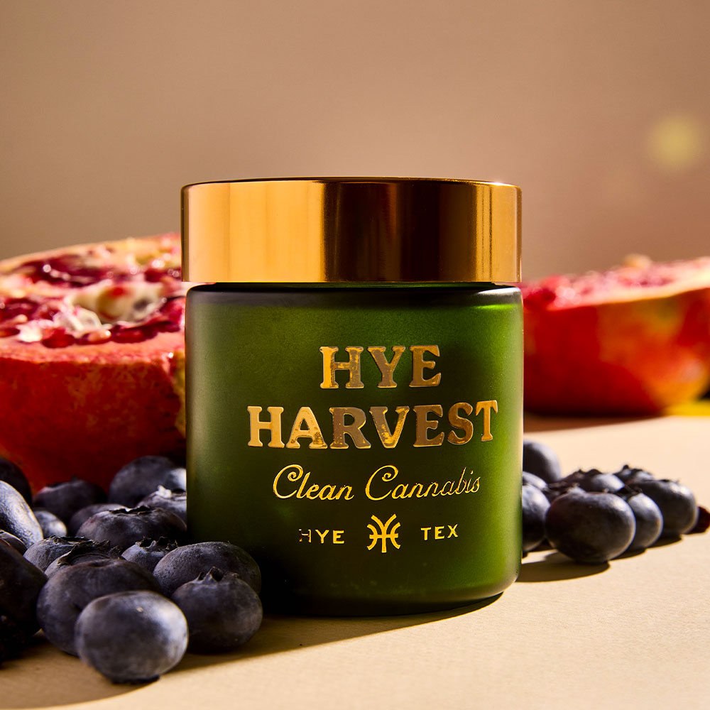A green glass jar with a gold lid featuring gold text that reads "Hye Harvest Clean Cannabis Hye Tex" sitting on a beige background surrounded by fresh blueberries and halved pomegranates.
